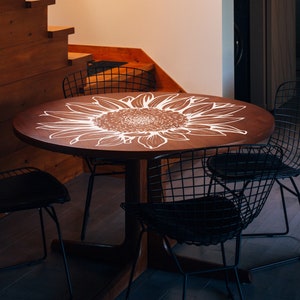 Puede incluir: Una mesa redonda de madera con un dise&ntilde;o de girasol blanco pintado en la superficie. La mesa est&aacute; rodeada por cuatro sillas de alambre negro.