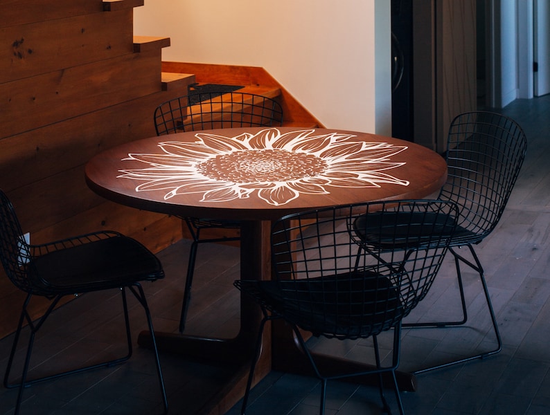 Puede incluir: Una mesa redonda de madera con un dise&ntilde;o de girasol blanco pintado en la superficie. La mesa est&aacute; rodeada por cuatro sillas de alambre negro.