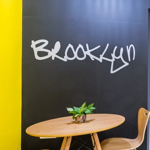 May include: A wooden table with a green plant in a pot sits in front of a black wall with the word "Brooklyn" written in white paint.