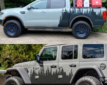 Op de afbeelding: Er worden twee voertuigen getoond: een lichtgrijze pick-up truck en een grijze Jeep Wrangler Rubicon. Beide voertuigen hebben zwarte bos-thema stickers op de zijkanten. De truck heeft blauwe wielen en een bedrek met rode en witte containers. De Jeep heeft zwarte wielen.