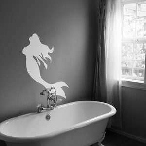 May include: A white silhouette of a mermaid with flowing hair is on a gray wall above a white bathtub with a silver faucet. The bathtub is in a bathroom with a white window and white curtains.