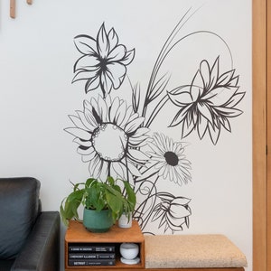 Op de afbeelding: Zwart-witte bloemendecoratie voor de muur met een zonnebloem, een madeliefje en andere bloemen. De decoratie is op een witte muur met een houten plank en een plant in een pot.