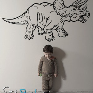 K&ouml;nnte beinhalten: Eine Schwarzwei&szlig;-Zeichnung eines Triceratops-Dinosauriers an einer wei&szlig;en Wand. Ein kleines Kind steht vor dem Dinosaurier und h&auml;lt einen gr&uuml;nen Ball.