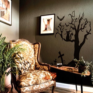 May include: A vintage-style armchair with floral upholstery sits in a room with a dark green wall. A black silhouette of a tree with bats and a cat is on the wall. The room is decorated with a framed picture of a staircase.