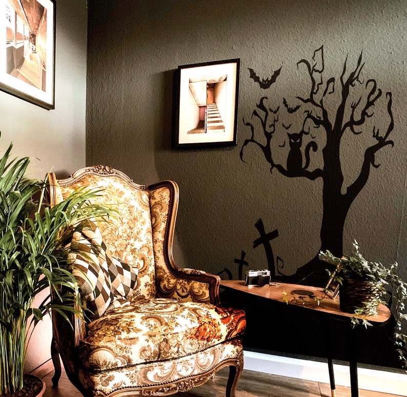 May include: A vintage-style armchair with floral upholstery sits in a room with a dark green wall. A black silhouette of a tree with bats and a cat is on the wall. The room is decorated with a framed picture of a staircase.