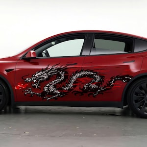 May include: A red Tesla Model Y with black wheels and a black dragon decal on the side. The dragon is black and white with red accents, and it stretches across the car's side.