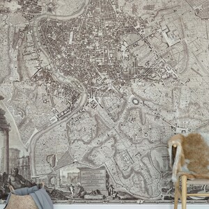 May include: A vintage map of Rome, Italy, printed in black and white. The map shows the city's streets, buildings, and landmarks. The map is framed and hanging on a wall. A wicker basket and a chair with a faux fur cushion are in the foreground.