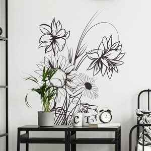 Op de afbeelding: Zwart-witte bloemendecoratie voor de muur met een zonnebloem, een madeliefje en andere bloemen. De decoratie is op een witte muur met een zwart rek en een plant in een pot ervoor.