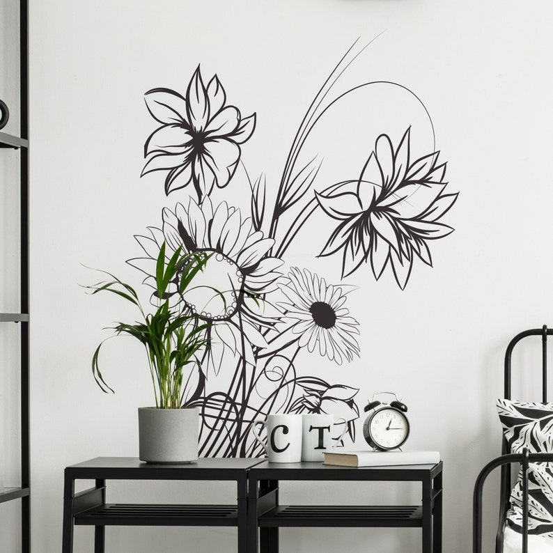 Op de afbeelding: Zwart-witte bloemendecoratie voor de muur met een zonnebloem, een madeliefje en andere bloemen. De decoratie is op een witte muur met een zwart rek en een plant in een pot ervoor.
