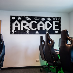 May include: A white wall with a black and white pixelated sign that says "ARCADE" with various pixelated images of video game characters and objects. Three black gaming chairs are in front of the wall.