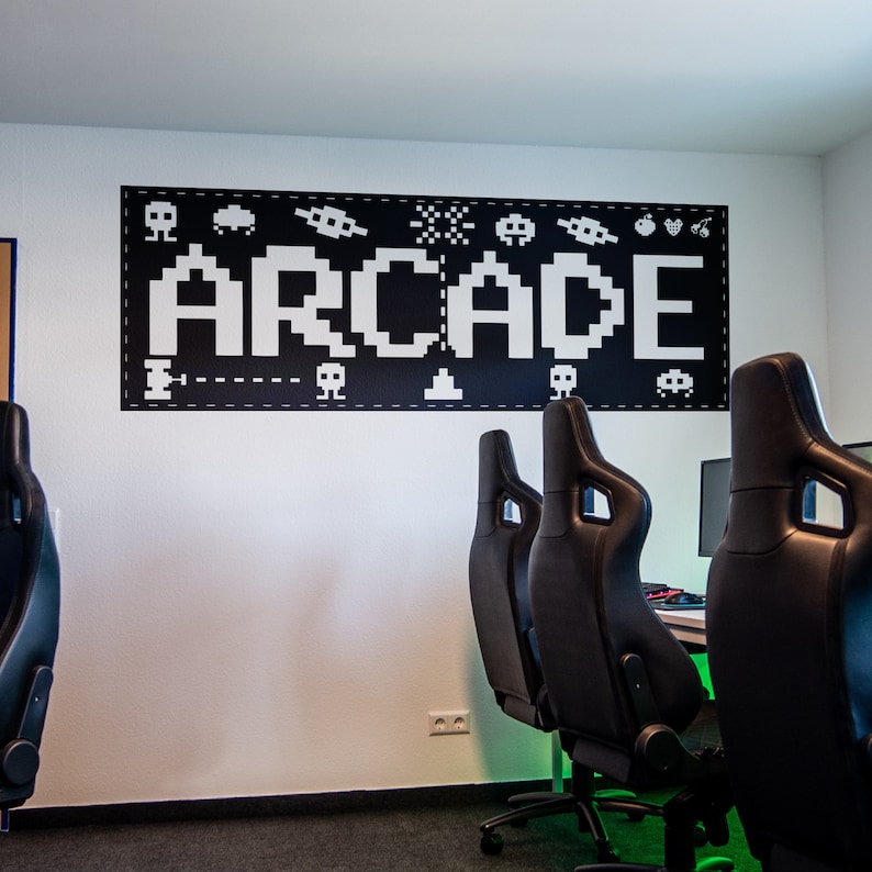 May include: A white wall with a black and white pixelated sign that says "ARCADE" with various pixelated images of video game characters and objects. Three black gaming chairs are in front of the wall.