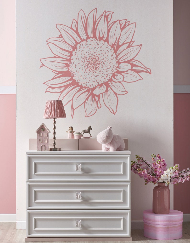 Puede incluir: Una c&oacute;moda blanca con tres cajones y pomos con forma de mariposa. La c&oacute;moda est&aacute; decorada con una calcoman&iacute;a de pared de girasol rosa y blanco. Una figura de hipop&oacute;tamo rosa se sienta encima de la c&oacute;moda.