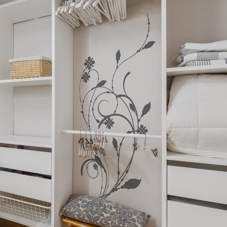 May include: White closet with shelves and a hanging rod. The closet has a decorative vinyl sticker of a gray floral design on the back wall. The closet is organized with folded clothes, a wicker basket, and a decorative pillow.