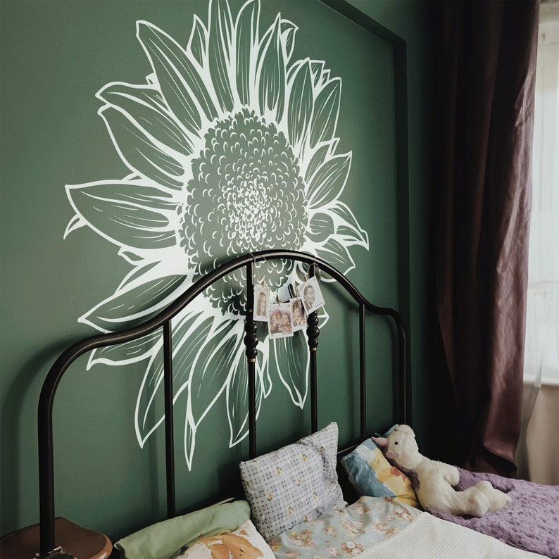 Puede incluir: Una cama con un marco de metal negro y un dise&ntilde;o de girasol blanco pintado en la pared detr&aacute;s de ella. La cama est&aacute; hecha con ropa de cama blanca y tiene una manta de tiro morada al pie de la cama. Hay un animal de peluche blanco en la cama.