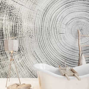 May include: A monochrome tree ring design adorns the wall behind a white bathtub. A wooden ladder leans against the wall. A floor lamp with a beige shade stands to the left of the tub. A wooden tray with glasses sits on the tub.