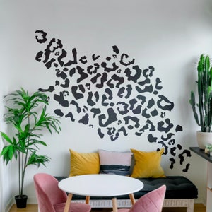 May include: A white wall with a black and white leopard print design. Two chairs with yellow and blue cushions are in front of a white table. A potted plant is in the corner of the room.