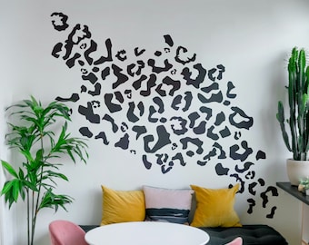 May include: A white wall with a black and white leopard print design. Two chairs with yellow and blue cushions are in front of a white table. A potted plant is in the corner of the room.