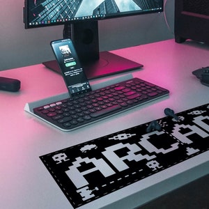 May include: A black and white pixelated "Arcade" design on a black mousepad. The mousepad is on a pink and white desk with a keyboard, a smartphone, and a computer monitor.