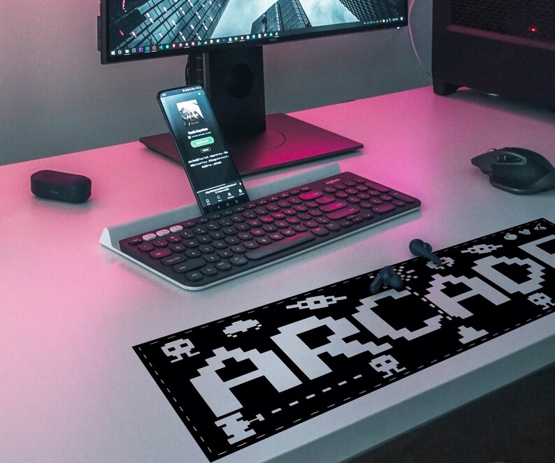 May include: A black and white pixelated "Arcade" design on a black mousepad. The mousepad is on a pink and white desk with a keyboard, a smartphone, and a computer monitor.