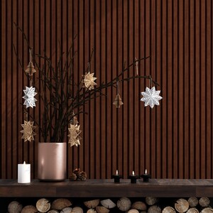 May include: A wooden wall with a brown branch with white and gold paper star ornaments hanging from it. A copper vase holds the branch. Three small candles are on a wooden shelf in front of the wall. A pile of logs is below the shelf.