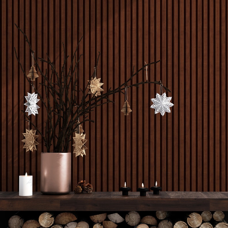 May include: A wooden wall with a brown branch with white and gold paper star ornaments hanging from it. A copper vase holds the branch. Three small candles are on a wooden shelf in front of the wall. A pile of logs is below the shelf.