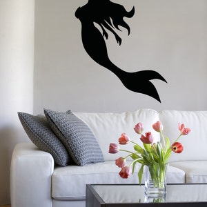 May include: A white couch with two gray pillows and a vase of pink tulips on a glass coffee table. A black silhouette of a mermaid is on the wall behind the couch.