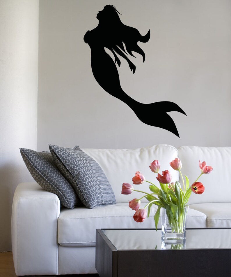 May include: A white couch with two gray pillows and a vase of pink tulips on a glass coffee table. A black silhouette of a mermaid is on the wall behind the couch.