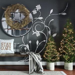 May include: A gray wall with a white floral design, a round mirror with a gold wreath, a bench with a white pillow that says "Stay Cozy", a black and white throw blanket, and two small Christmas trees with white lights.