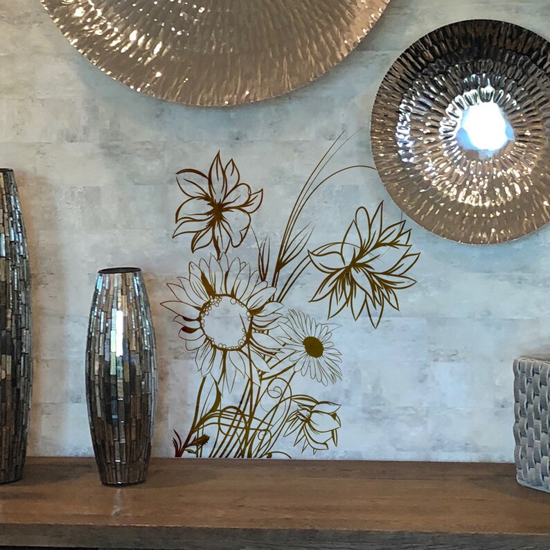 Op de afbeelding: Een decoratieve muursticker met een boeket gestileerde bloemen in bruin tegen een lichtgrijze achtergrond. Twee zilveren decoratieve vazen staan op een houten plank onder de sticker.