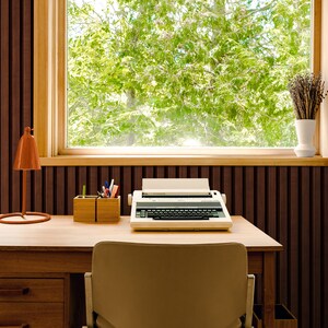 May include: A vintage typewriter sits on a wooden desk in front of a window with a view of green trees. The desk has a lamp and a pencil holder with pencils and pens.