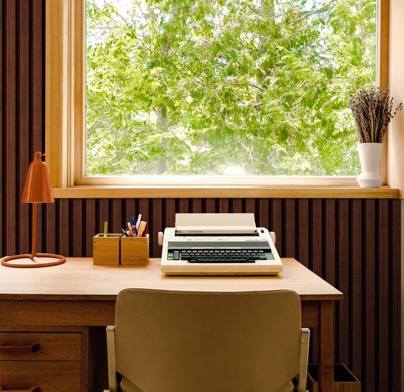 May include: A vintage typewriter sits on a wooden desk in front of a window with a view of green trees. The desk has a lamp and a pencil holder with pencils and pens.