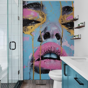May include: A bathroom with a large abstract painting of a woman's face with pink lips and gold accents on a blue background. The painting is above a white toilet and a blue vanity with a white countertop.