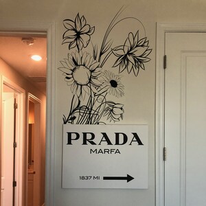 Op de afbeelding: Een wit bord met zwarte tekst dat "PRADA MARFA 1837 MI" aangeeft met een zwarte pijl die naar rechts wijst. Het bord is gemonteerd op een witte muur met een zwart bloemenontwerp.