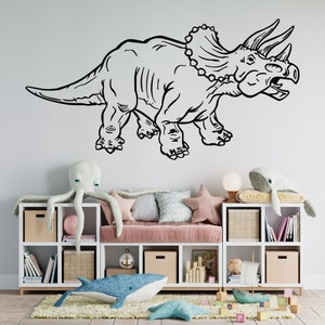 K&ouml;nnte beinhalten: Schwarz-wei&szlig;-Zeichnung eines Triceratops-Dinosauriers an einer wei&szlig;en Wand. Der Dinosaurier blickt nach rechts und hat drei H&ouml;rner auf dem Kopf. Der Dinosaurier befindet sich &uuml;ber einem wei&szlig;en B&uuml;cherregal mit einem rosa Kissen darauf. Auf dem Regal und auf dem Boden befinden sich Stofftiere.