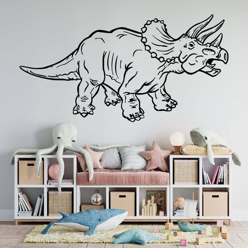 K&ouml;nnte beinhalten: Schwarz-wei&szlig;-Zeichnung eines Triceratops-Dinosauriers an einer wei&szlig;en Wand. Der Dinosaurier blickt nach rechts und hat drei H&ouml;rner auf dem Kopf. Der Dinosaurier befindet sich &uuml;ber einem wei&szlig;en B&uuml;cherregal mit einem rosa Kissen darauf. Auf dem Regal und auf dem Boden befinden sich Stofftiere.