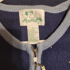 May include: A navy blue knit cardigan with light blue trim, a silver zipper, and a cat-shaped zipper pull. The interior label reads The Quacker Factory, size S, 54% ramie, 44% cotton, 2% other fibers, RN 95524, looped and stitched in Hong Kong.