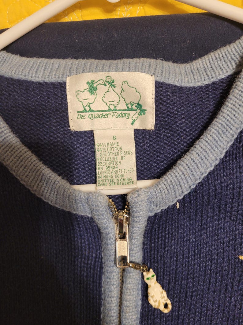 May include: A navy blue knit cardigan with light blue trim, a silver zipper, and a cat-shaped zipper pull. The interior label reads The Quacker Factory, size S, 54% ramie, 44% cotton, 2% other fibers, RN 95524, looped and stitched in Hong Kong.
