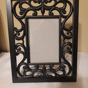 May include: Black rectangular picture frame with ornate scrollwork and a central opening for a photo. The frame has a matte finish and a neutral background.