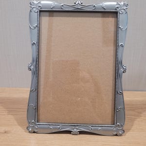 May include: A rectangular silver-colored picture frame featuring an ornate, curved border with intricate scrollwork details and small embedded clear stones. The frame has a beaded inner edge and a plain, light brown cardboard backing insert.