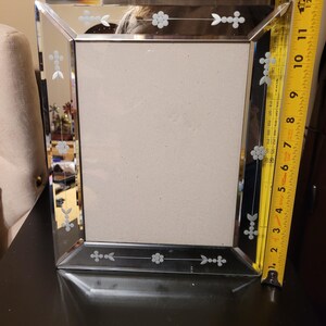 Puede incluir: Un marco de fotos rectangular espejado con dise&ntilde;os florales y de flechas grabados. El marco mide aproximadamente 25 cm de alto y 20 cm de ancho. El marco est&aacute; sobre una superficie oscura.