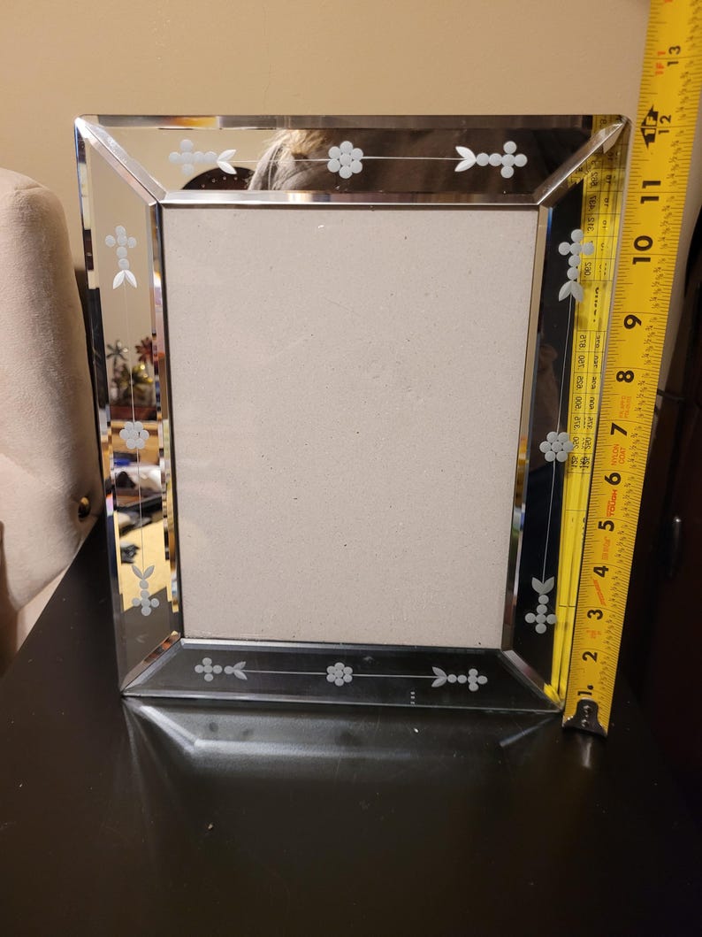 Puede incluir: Un marco de fotos rectangular espejado con dise&ntilde;os florales y de flechas grabados. El marco mide aproximadamente 25 cm de alto y 20 cm de ancho. El marco est&aacute; sobre una superficie oscura.