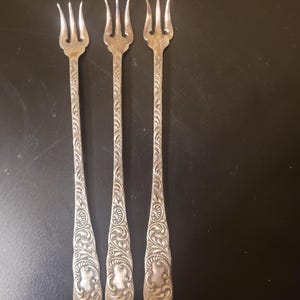 May include: Three silver-toned metal cocktail forks featuring a three-tined head and a long, slender handle. Each handle is decorated with an intricate, embossed scrollwork pattern that extends from the base up to the neck of the fork.