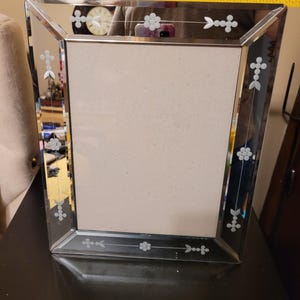 Puede incluir: Un marco de fotos rectangular espejado con dise&ntilde;os florales grabados. El marco mide aproximadamente 20 cm de ancho. El marco tiene un borde biselado y un respaldo de color neutro.