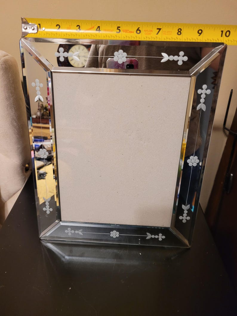 Puede incluir: Un marco de fotos rectangular espejado con dise&ntilde;os florales grabados. El marco mide aproximadamente 20 cm de ancho. El marco tiene un borde biselado y un respaldo de color neutro.