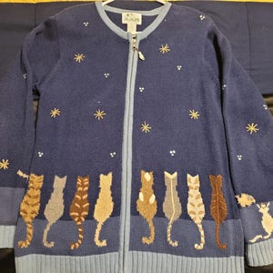 May include: A navy blue zip-up cardigan featuring a row of embroidered cats along the bottom hem. The sweater is decorated with small white dots and gold star-shaped embroidery across the chest and sleeves, with light blue ribbed trim.