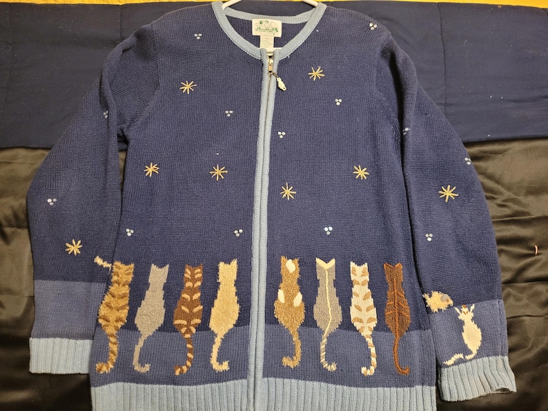 May include: A navy blue zip-up cardigan featuring a row of embroidered cats along the bottom hem. The sweater is decorated with small white dots and gold star-shaped embroidery across the chest and sleeves, with light blue ribbed trim.