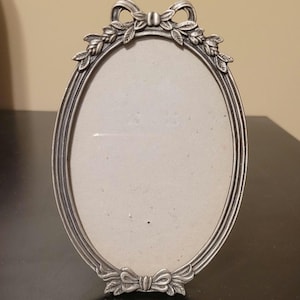 May include: Silver-toned oval picture frame with a decorative bow and leaf design at the top and bottom. The frame has a smooth, polished finish and is designed to hold a photograph or artwork. The frame is standing upright on a dark surface.