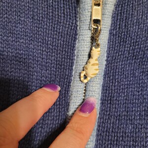May include: A dark blue knit zip-up jacket featuring a light blue trim along the zipper line. The metal zipper pull is adorned with a small, cream-colored cat charm with brown stripes, attached by a small lobster clasp to the zipper slider.