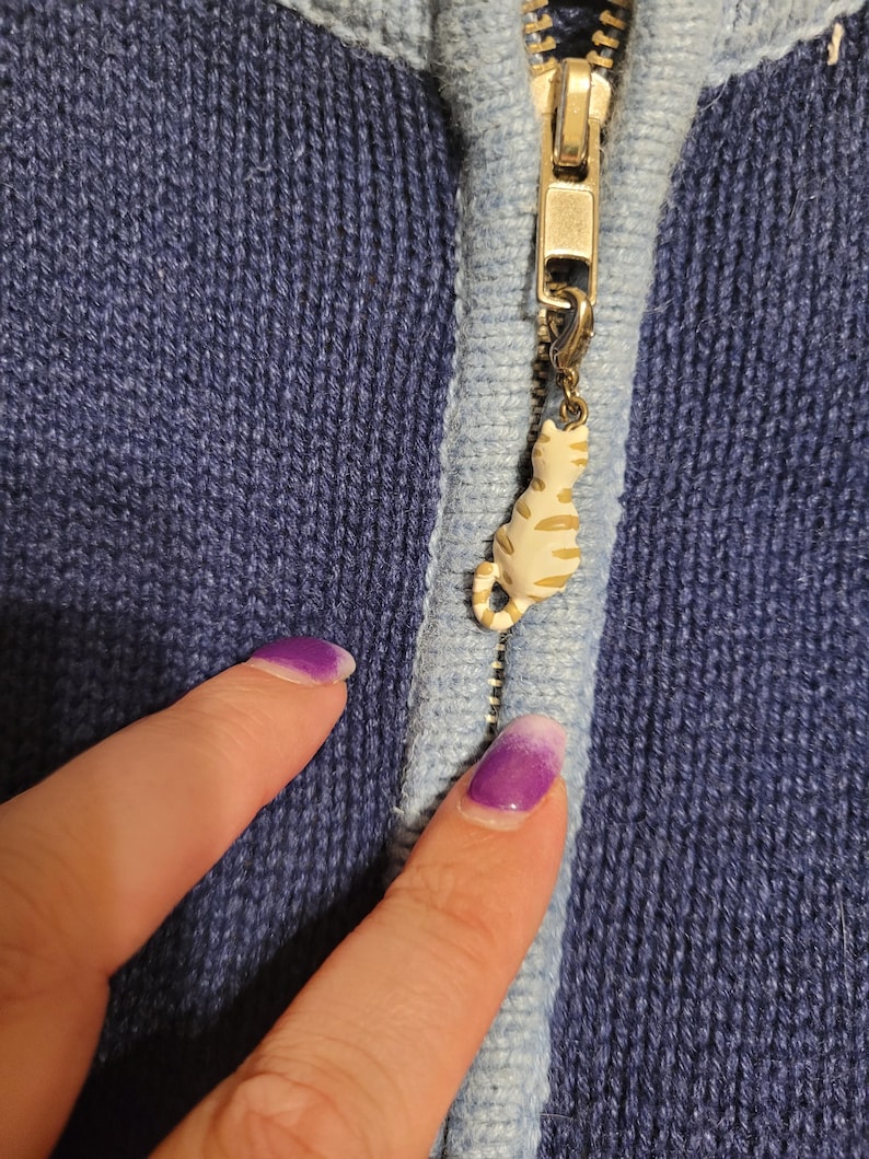 May include: A dark blue knit zip-up jacket featuring a light blue trim along the zipper line. The metal zipper pull is adorned with a small, cream-colored cat charm with brown stripes, attached by a small lobster clasp to the zipper slider.