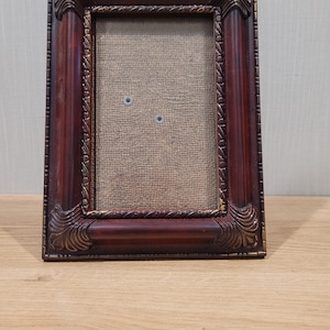May include: A rectangular picture frame with a dark reddish-brown finish and ornate carved corner details. The frame features a textured, light brown fabric-covered backing insert with two small circular metal pins visible in the center area.